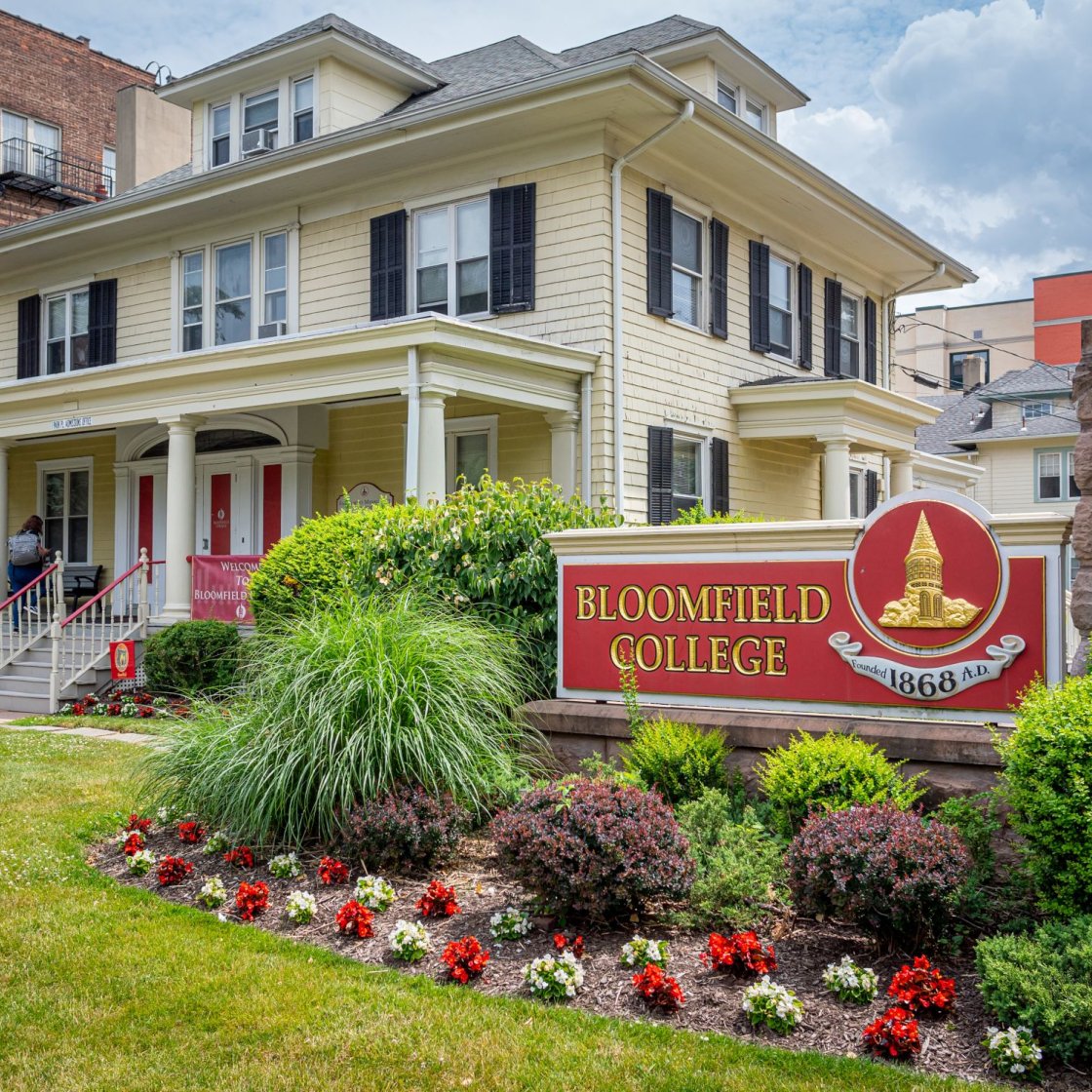A yellow house with a Bloomfield College sign surrounded by green bushes.