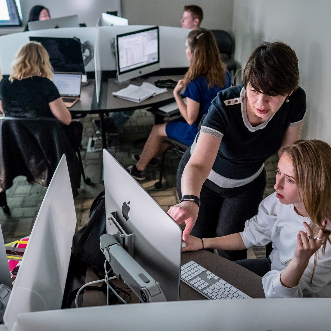 Teacher pointing on a computer to another student, while a group of students are behind on their own computers.