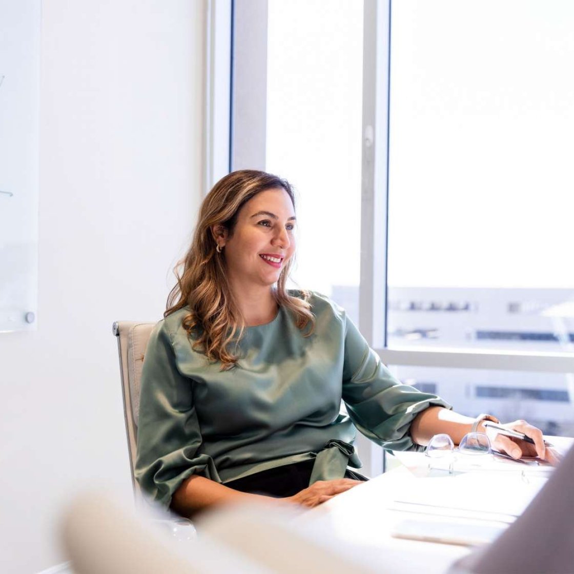 Woman in green blouse sits at desk with pen in hand speaking with a colleague off screen.
