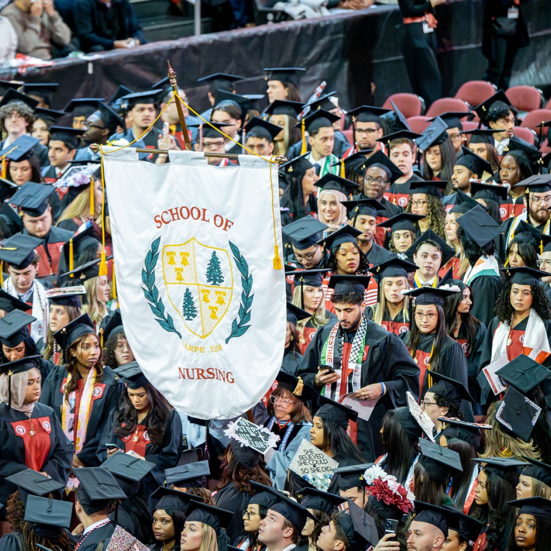 Students at commencement with their black cap and gowns, holding up a banner that says School of Nursing.