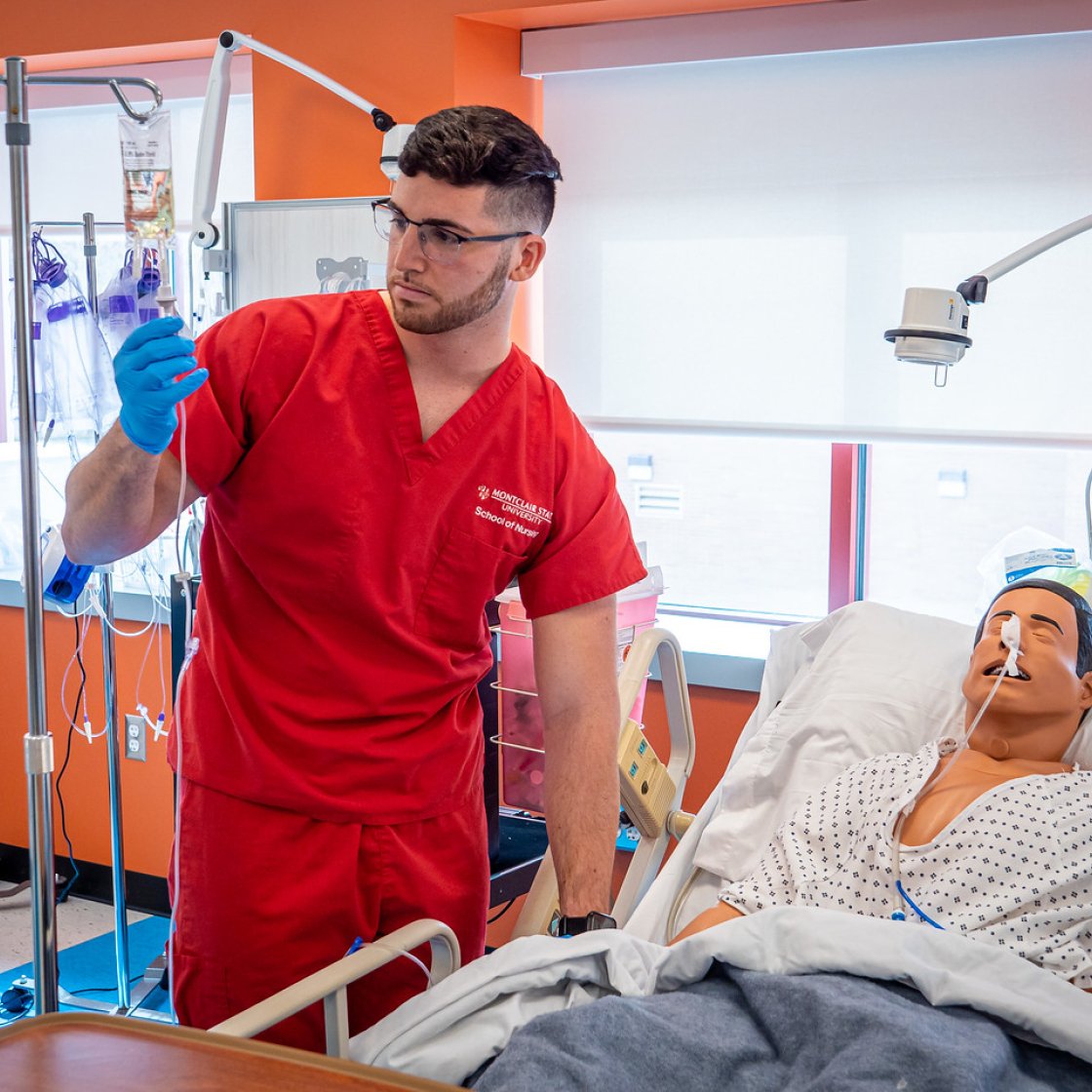 Nursing student in red scrubs checks the IV in a bag on a dummy.