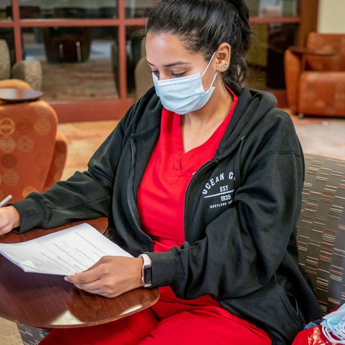 A student in red scrubs and a surgical mask completes her homework in a waiting room