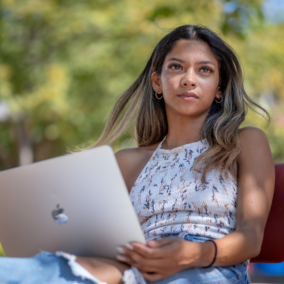 A student sits outside on a chair on a sunny day, holding an open laptop on their lap and studying in front of a backdrop of green, leafy trees.