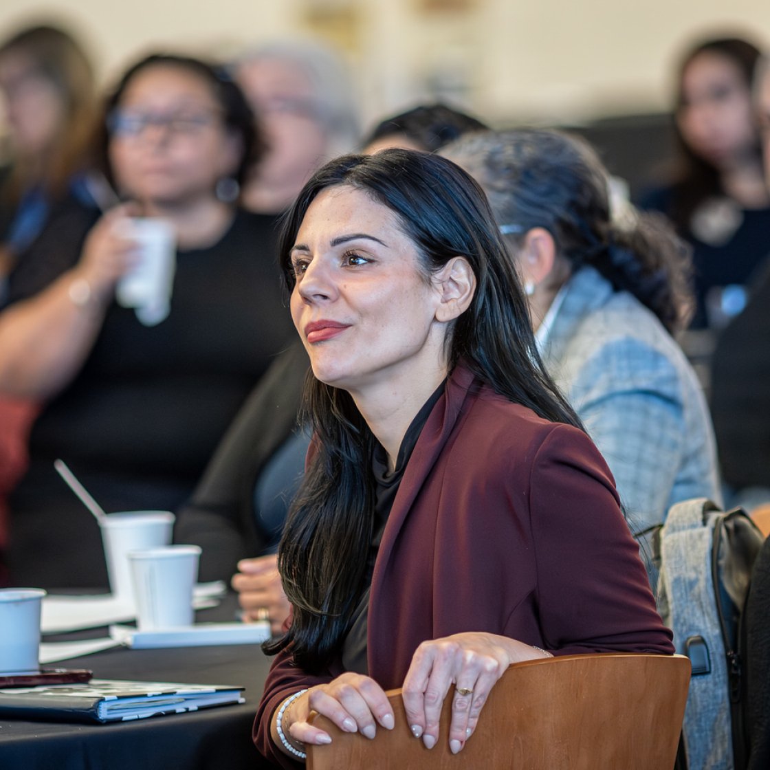 A person in a maroon blazer sits at a table during a conference event, listening attentively, with other attendees and coffee cups visible in the blurred background.