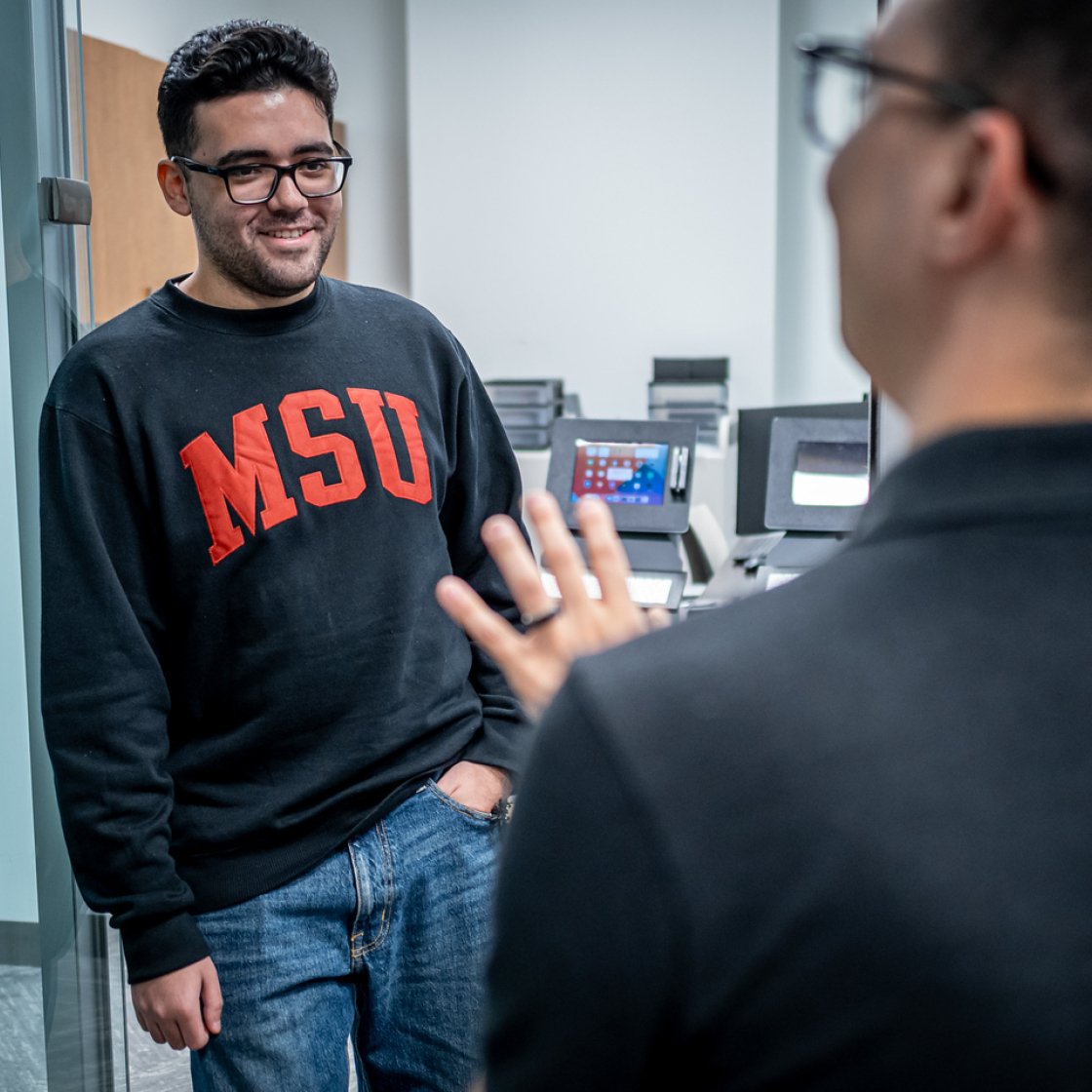 A student wearing a Montclair sweatshirt stands near a glass wall in an office, listening to another person in the foreground who is speaking with one hand raised.