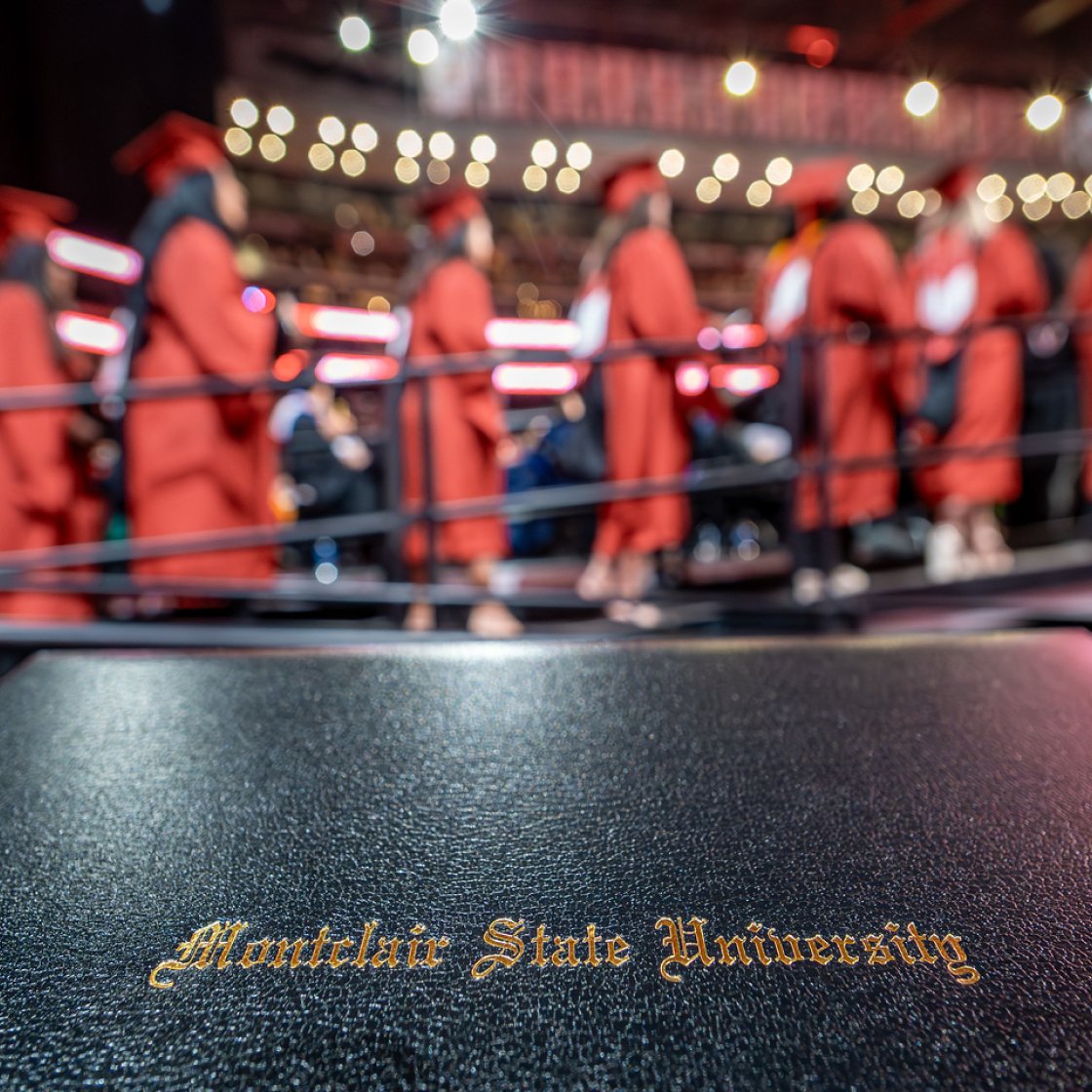 Graduates in red cap and gowns in the background while there is a diploma in front with the words Montclair State University.