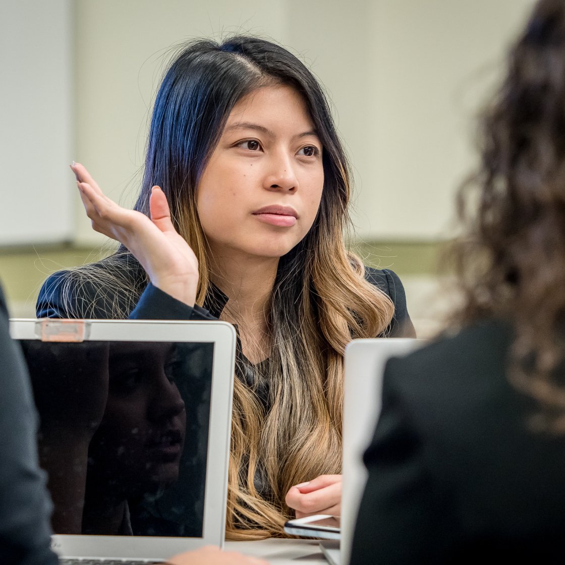 A student speaks during a small group discussion around laptops in a classroom, gesturing with one hand as classmates listen.