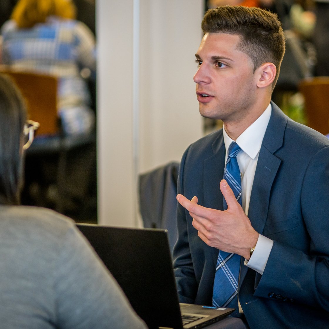 A man in a suit and blue tie gesturing and explaining to another person.