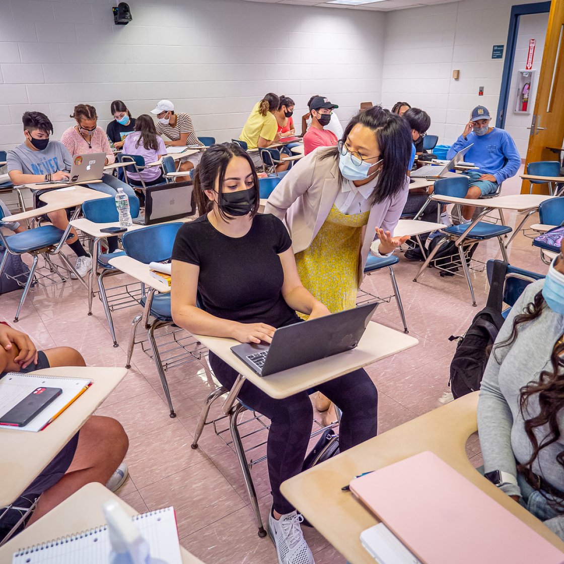 Teacher is explaining something to a student sitting down with her laptop in front of her.