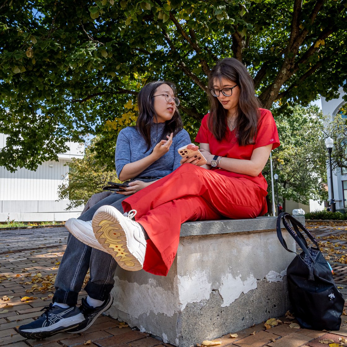 Two students siting on a stone bench as they talk.