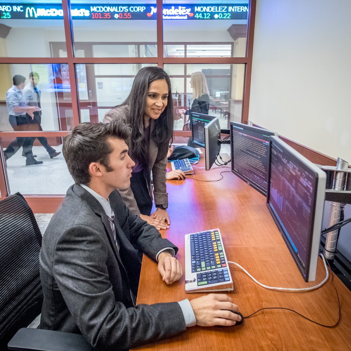 Students work at a multi‑monitor trading station, analyzing financial data in a glass‑walled business lab.