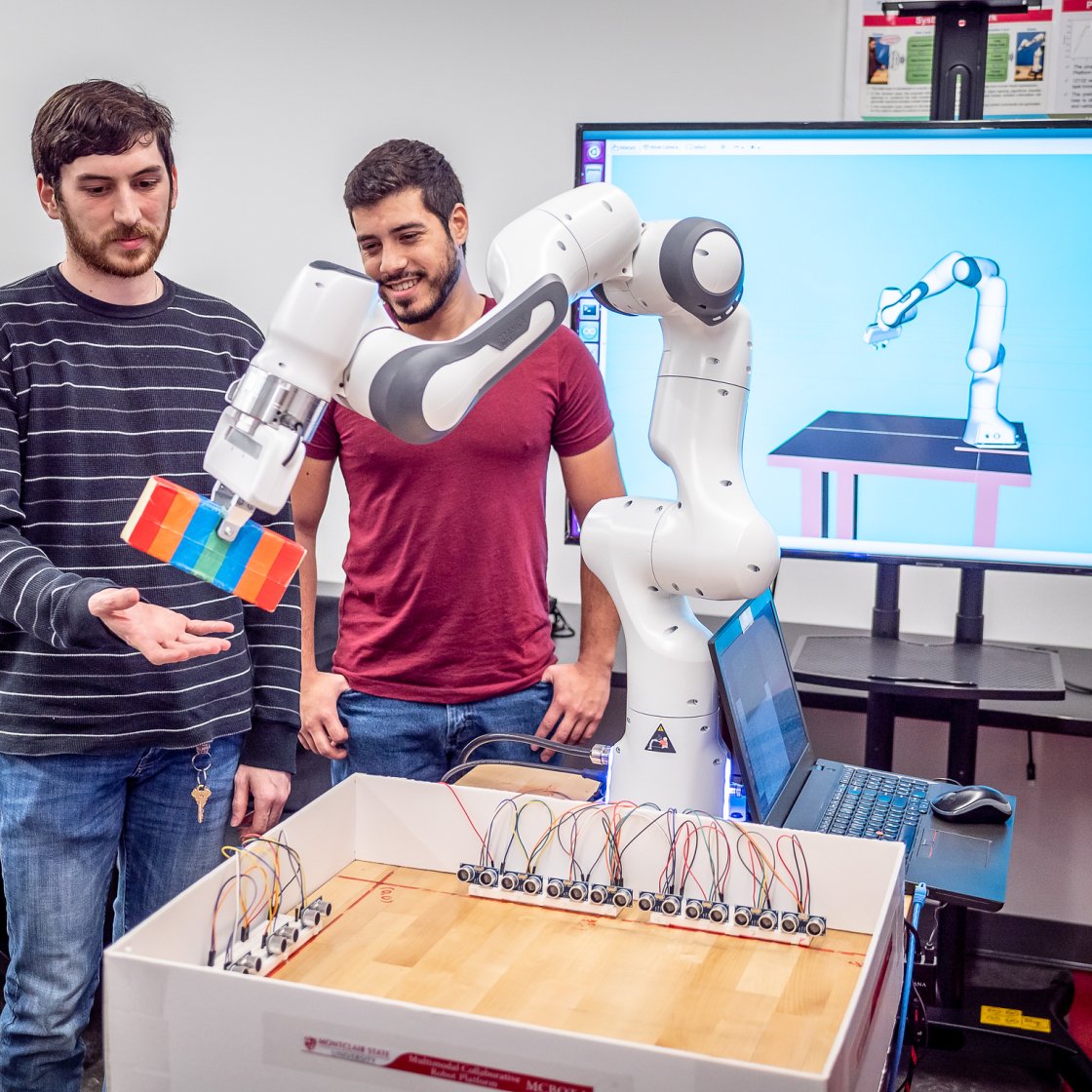 Two students work with a white robotic arm that is stacking colorful blocks, while a computer screen behind them shows a 3D simulation of the same robot.