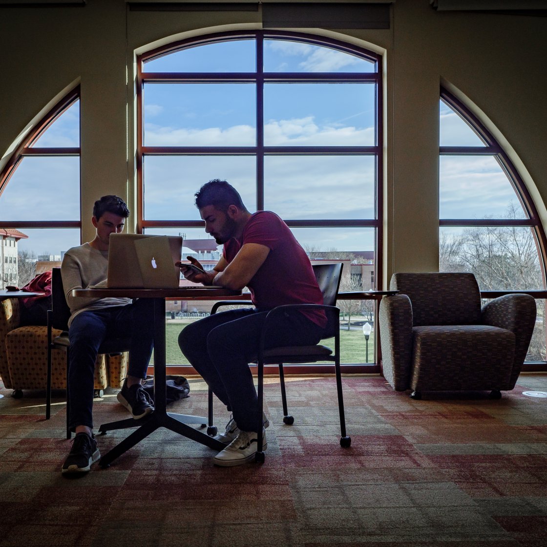 Two students study together at a small table in front of a large arched window overlooking campus, working on a laptop in a bright lounge area.