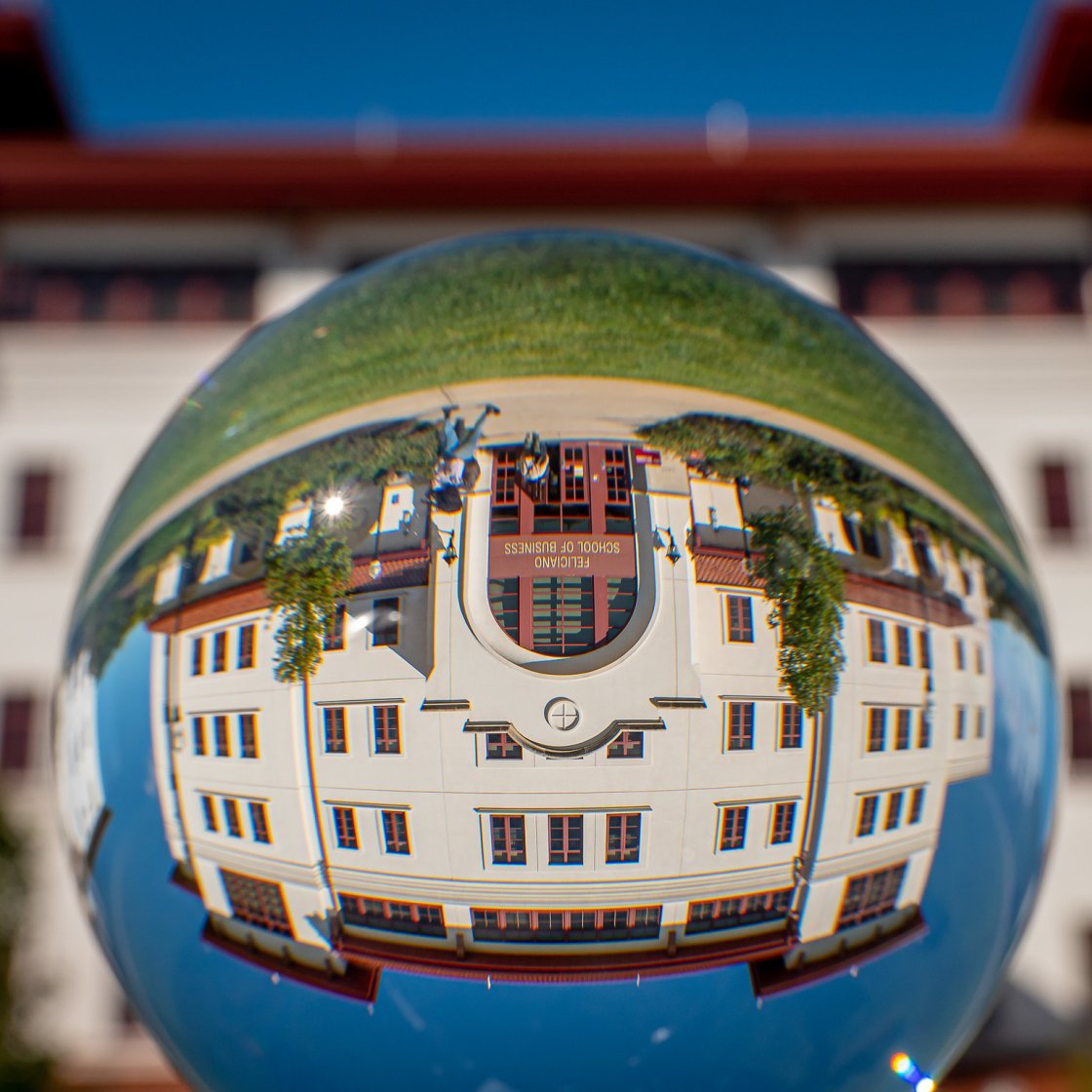 A glass sphere reflects an upside‑down view of the Feliciano School of Business building against a bright blue sky.