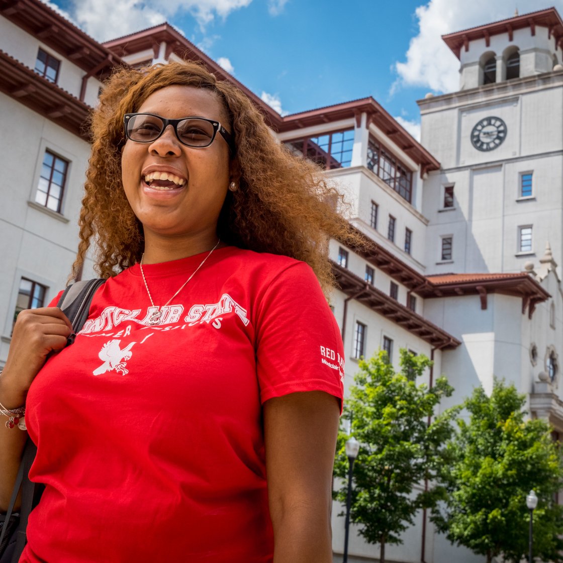A student wearing a red Montclair Red Hawk t-shirt smiles while walking on campus.