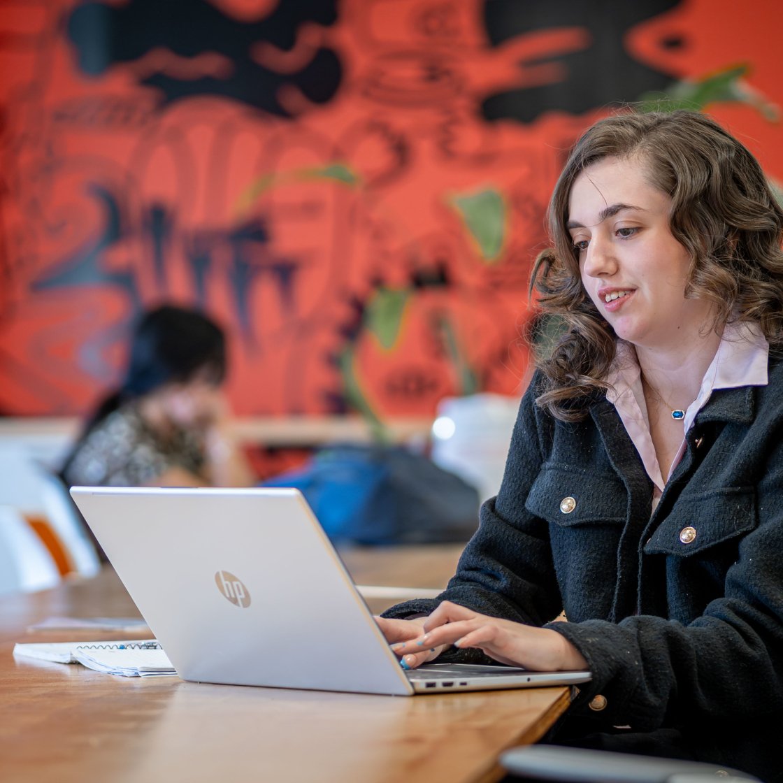 A student works on a laptop at a long table in a colorful study space with a bright mural on the wall behind her.