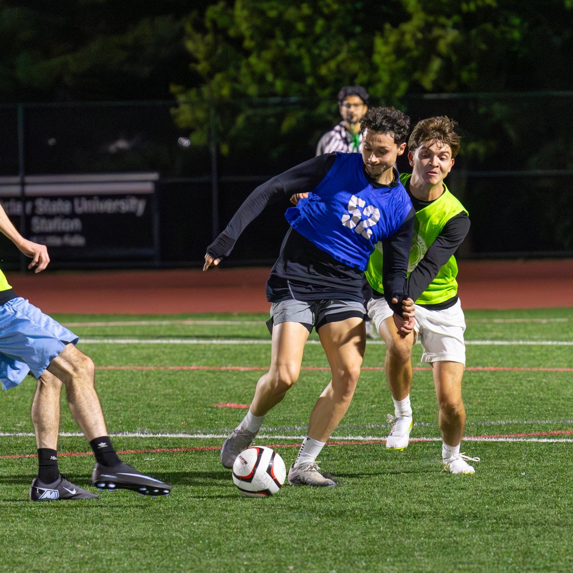 Three students on a pitch playing soccer kicking a soccer ball.