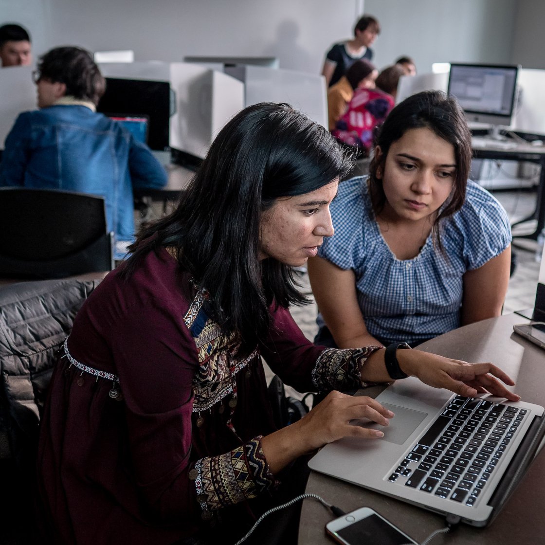Students collaborate on a laptop in a computer lab, with classmates working at other computers in the background.