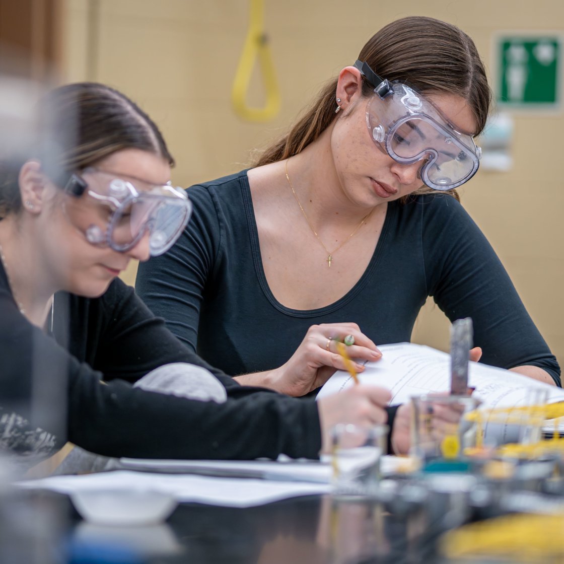 Two female students wearing safety goggles are taking notes.