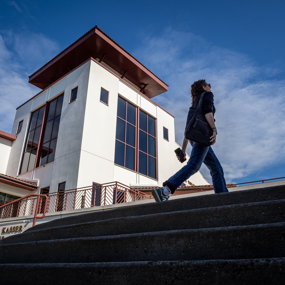 A person walks up the steps outside the Alexander Kasser Theater at Montclair State University under a bright blue sky.