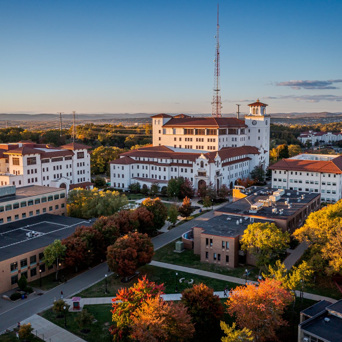 A scenic aerial view of Montclair State University’s campus, featuring academic buildings with red-tiled roofs, tree-lined walkways and autumn foliage.​