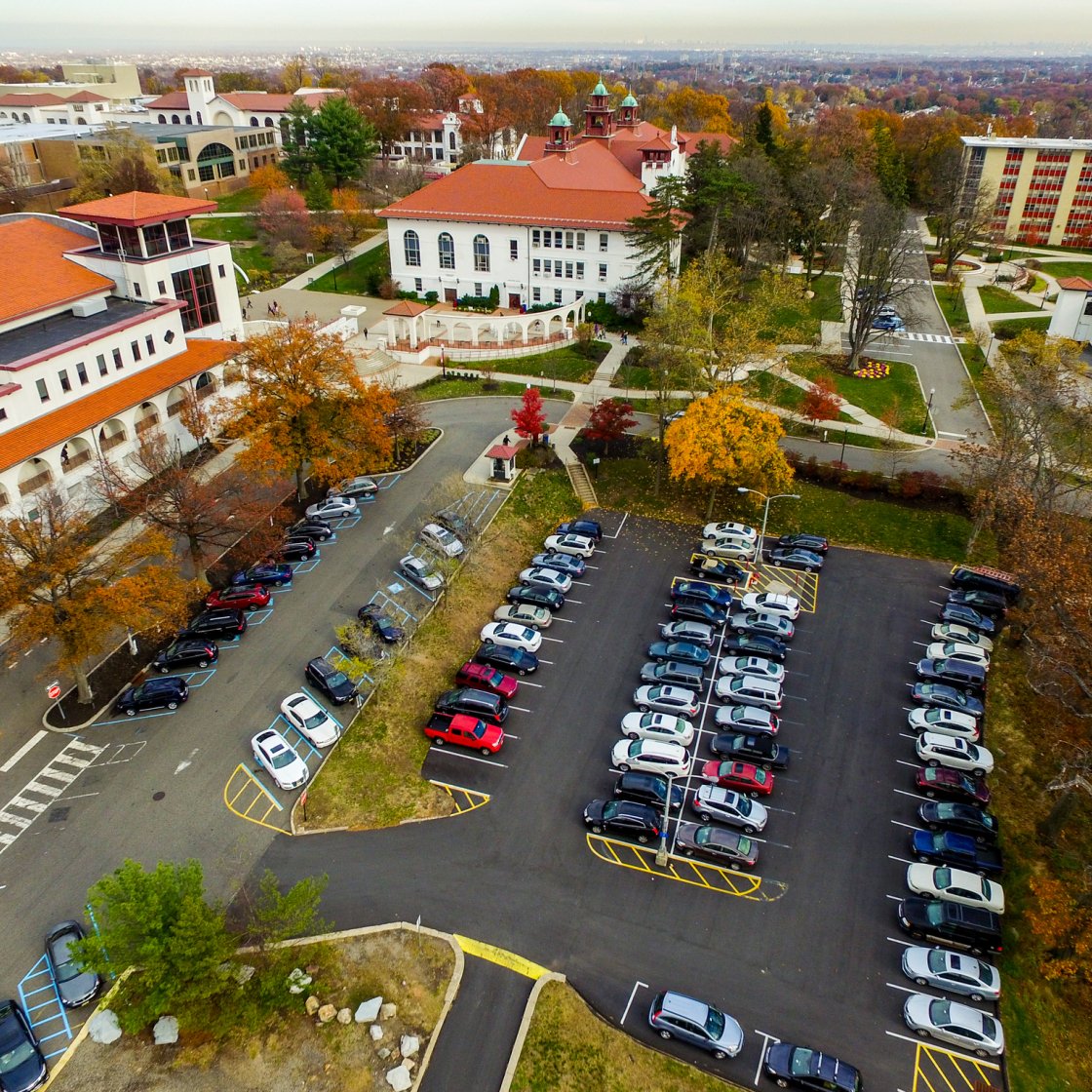 Aerial view of Montclair State University’s campus in autumn, showing red-roofed academic buildings, tree-lined walkways, and several full parking lots with rows of parked cars.