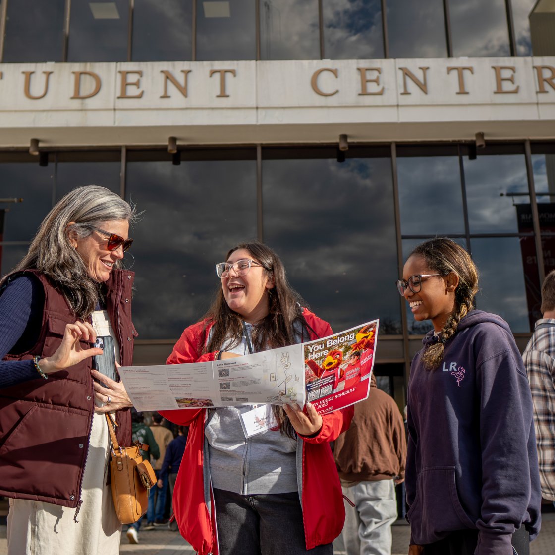 Three people stand in front of the Montclair State University Student Center, looking at a brochure together.