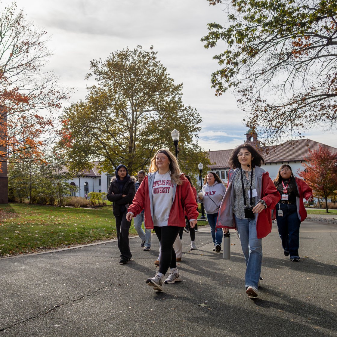 A group of students and a campus tour guide walk along a tree-lined path at Montclair State University in autumn.