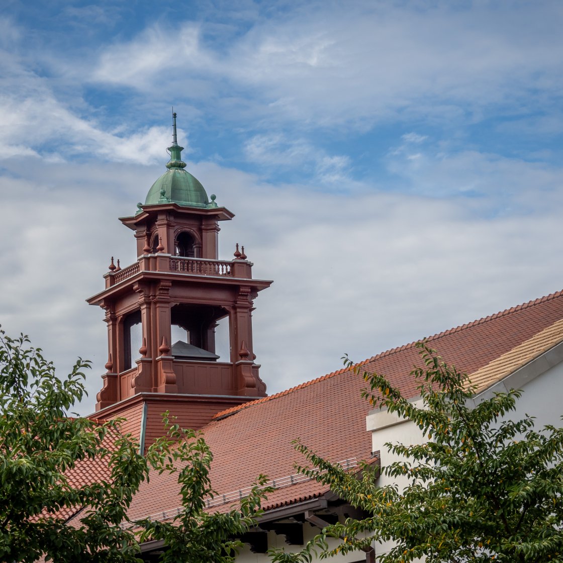 The image shows the bell tower of Cole Hall with a cloudy blue sky in the background and green tree leaves in the foreground.