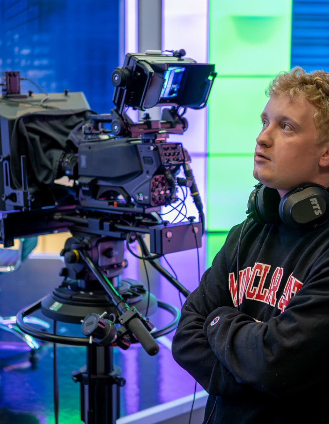 Brian Scott stands with arms crossed in a brightly lit TV studio, wearing a headset around their neck beside a large professional video camera and teleprompter.