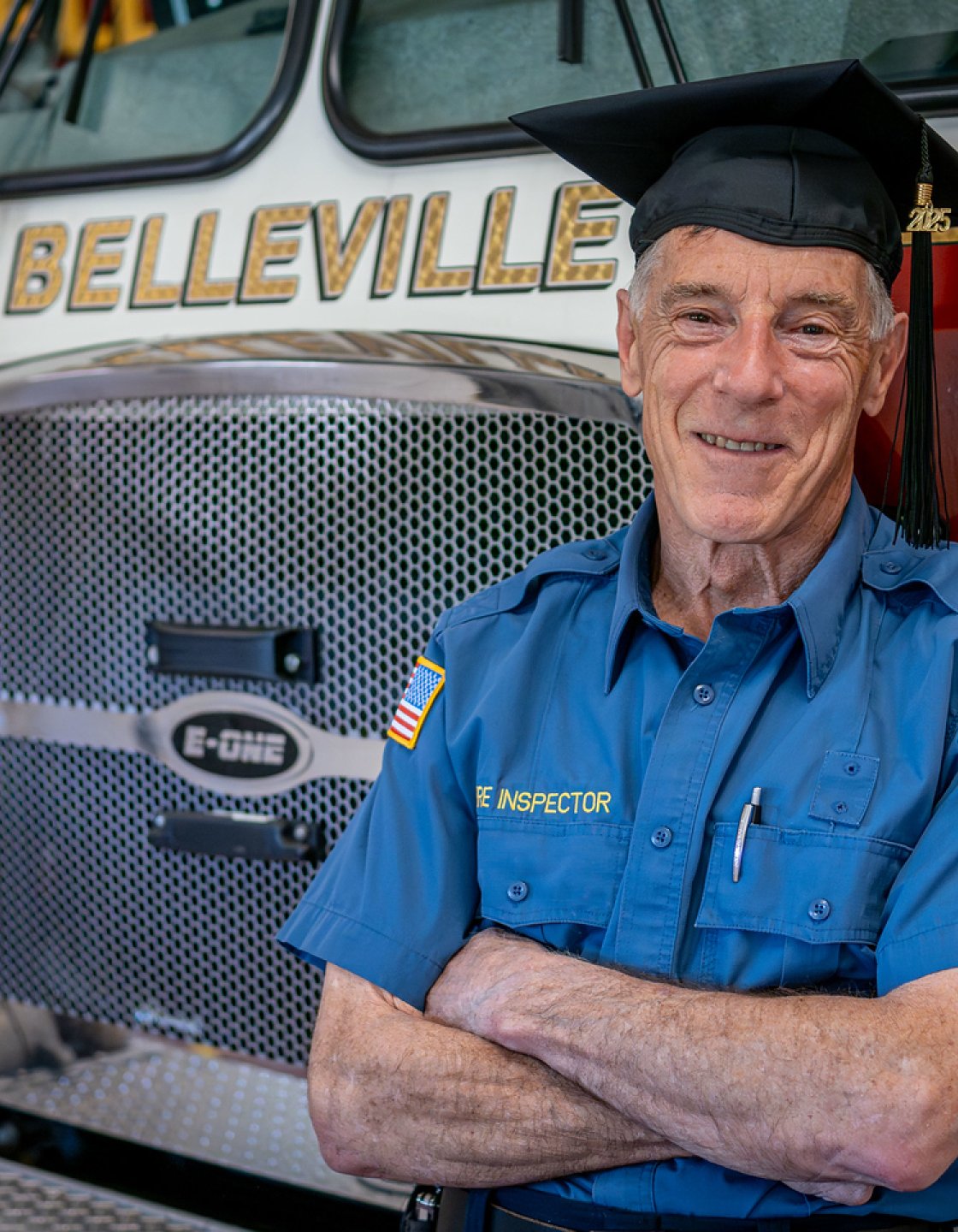 Ralph Castellano stands in front of a fire truck in the station, wearing his uniform and graduation cap.