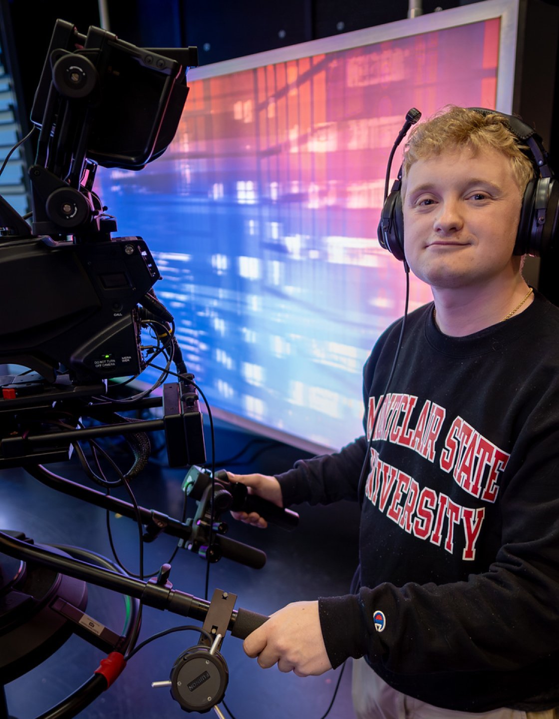 Brian Scott stands in a television studio behind a professional video camera, wearing a Montclair State University sweatshirt and a headset.