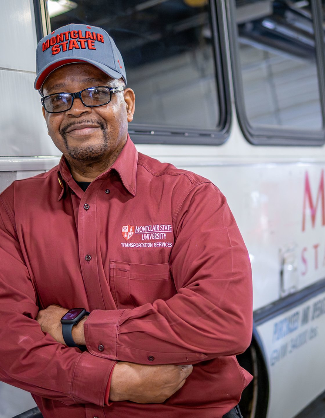 Frank Rivers, wearing a red Montclair Facilities uniform, stands in front of a campus shuttle bus.