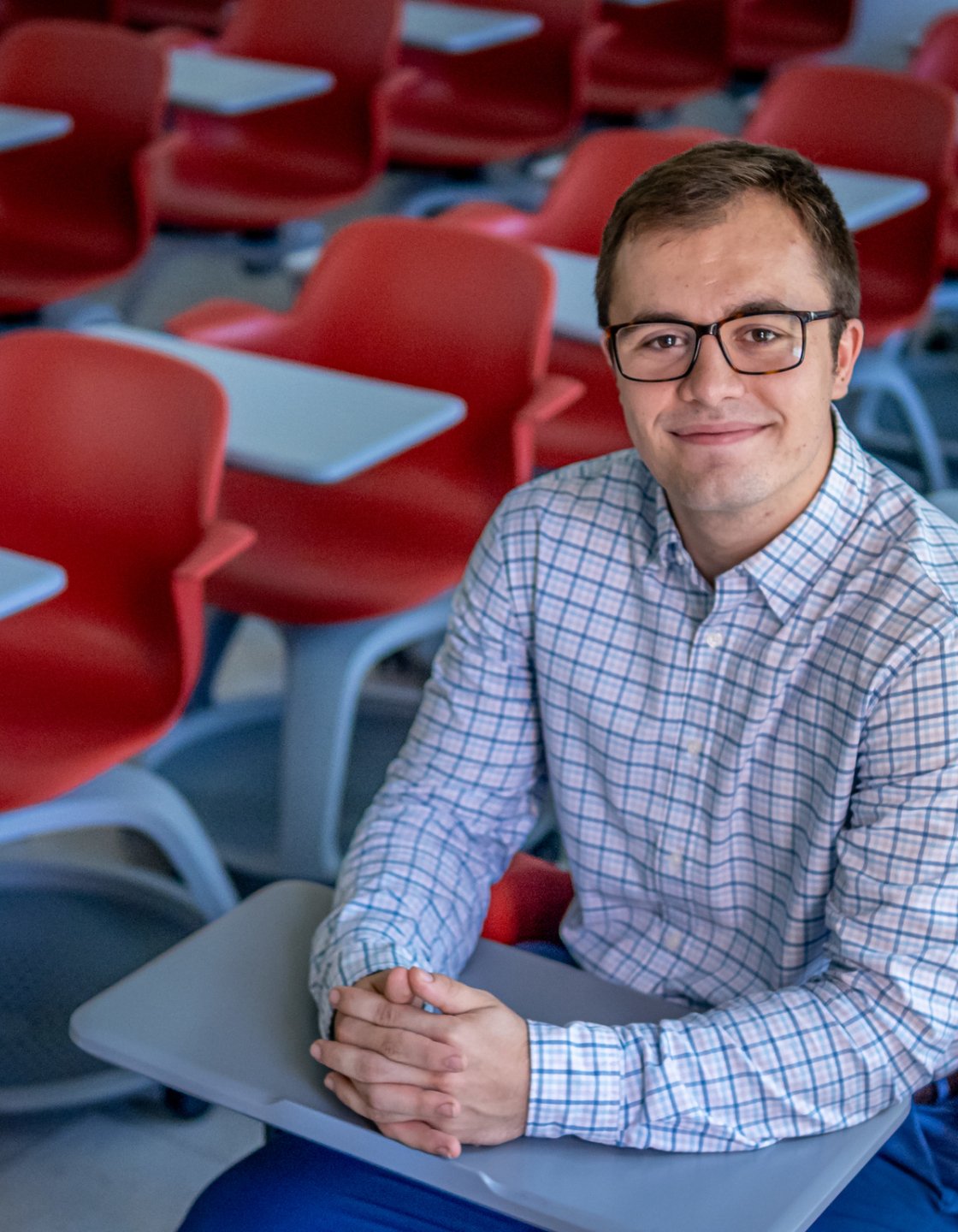 Jacob Roby sits in a classroom filled with red chairs and tablet desks.