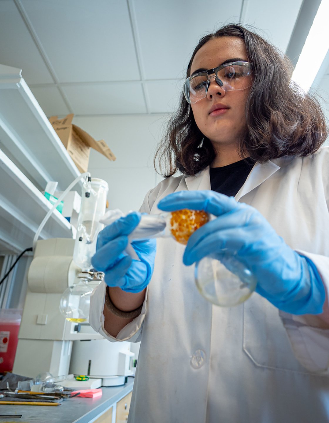 Rachel Gushikem, wearing a white lab coat and blue gloves, stands at a lab bench, holding a round-bottom flask and a clump of orange material wrapped in gauze, surrounded by shelves of equipment and bottles.