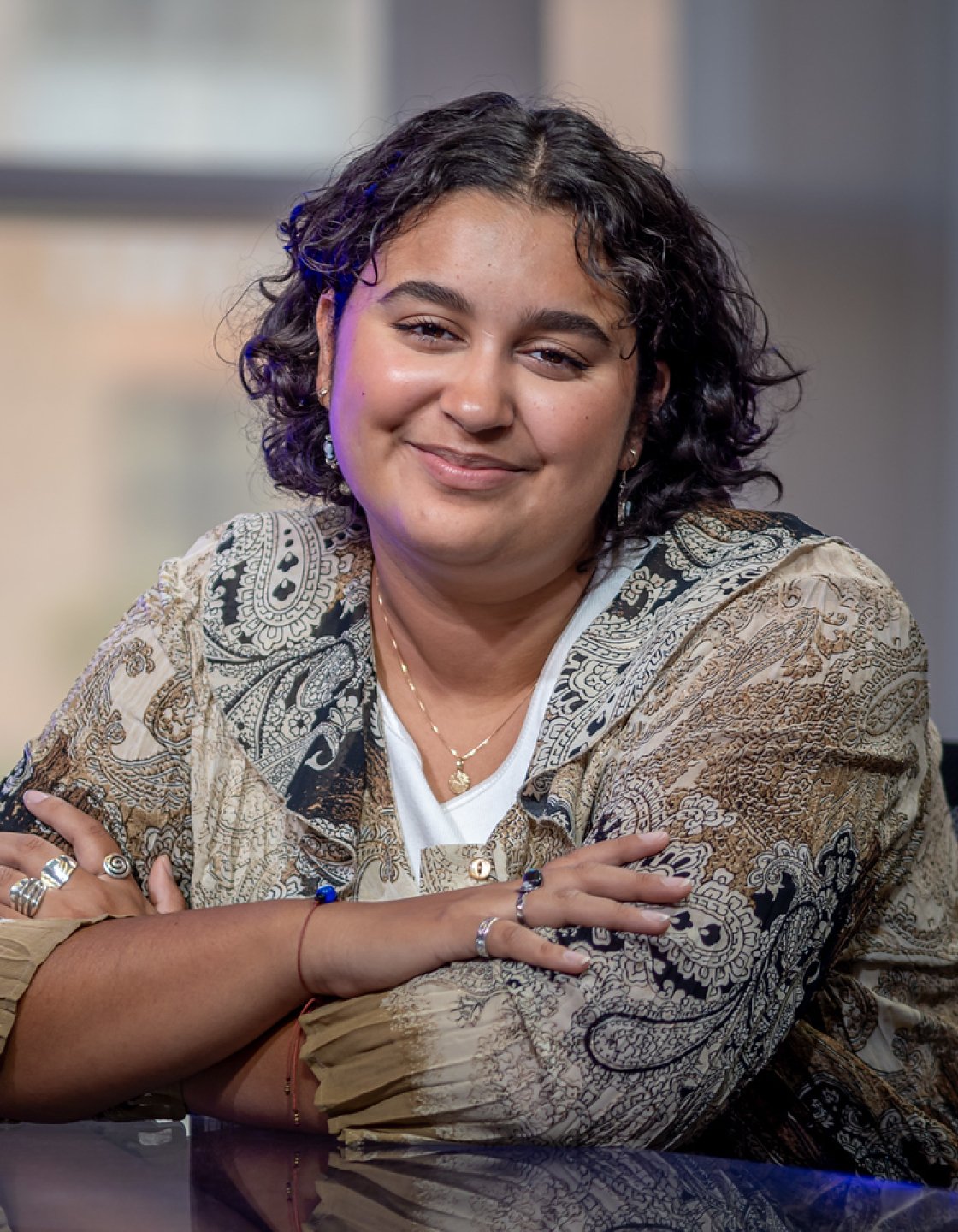 Journalism student Dani Mazariegos smiling with arms folded on a desk, seated in front of large windows in a bright, modern studio space.