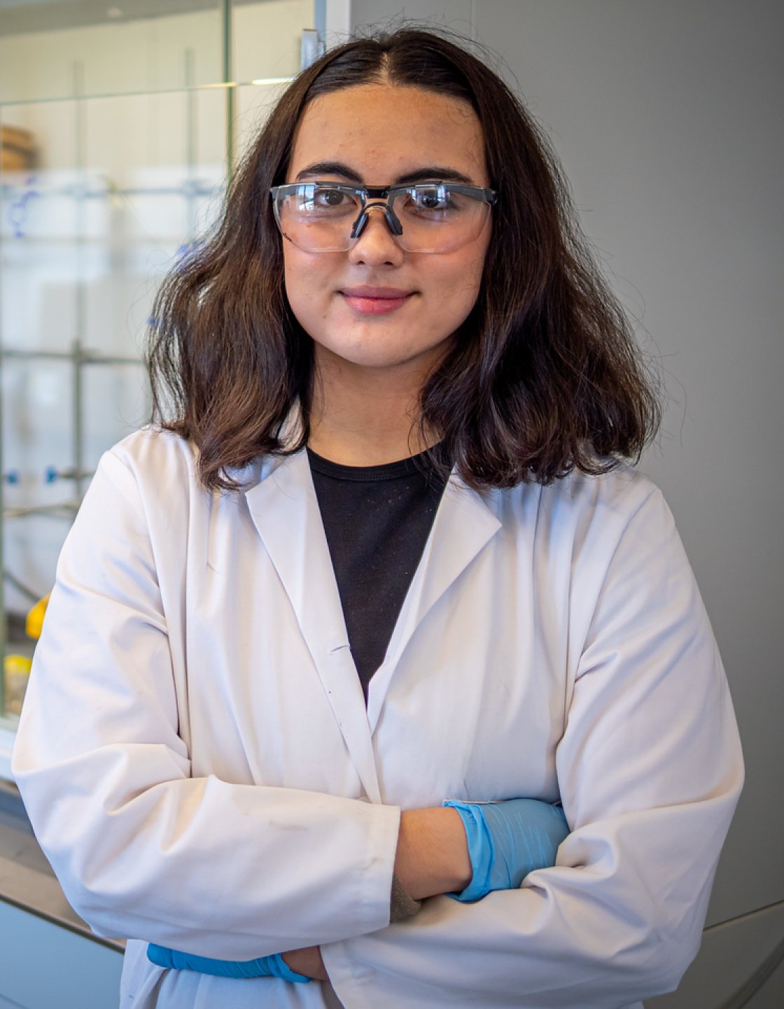 Student researcher Rachel Gushikem wearing safety goggles and a lab coat, standing with folded arms in front of laboratory equipment and a fume hood.