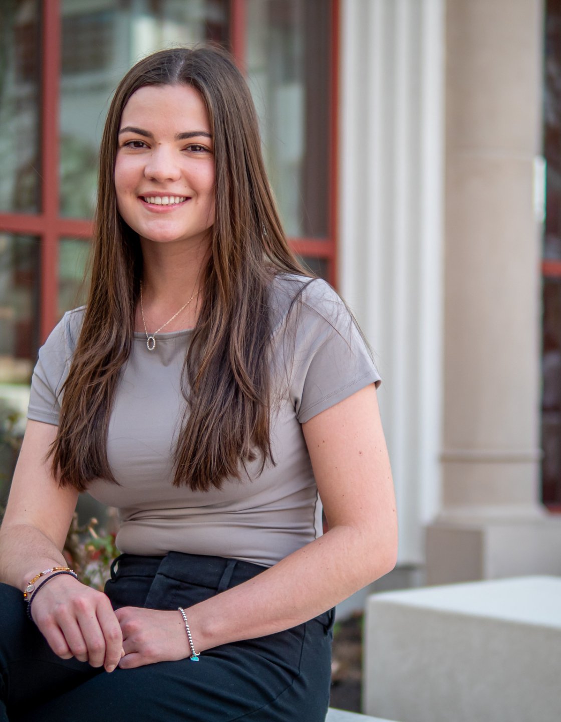 Madison Surgent sits outside a campus building, hands folded in her lap, with large windows and columns in the background.