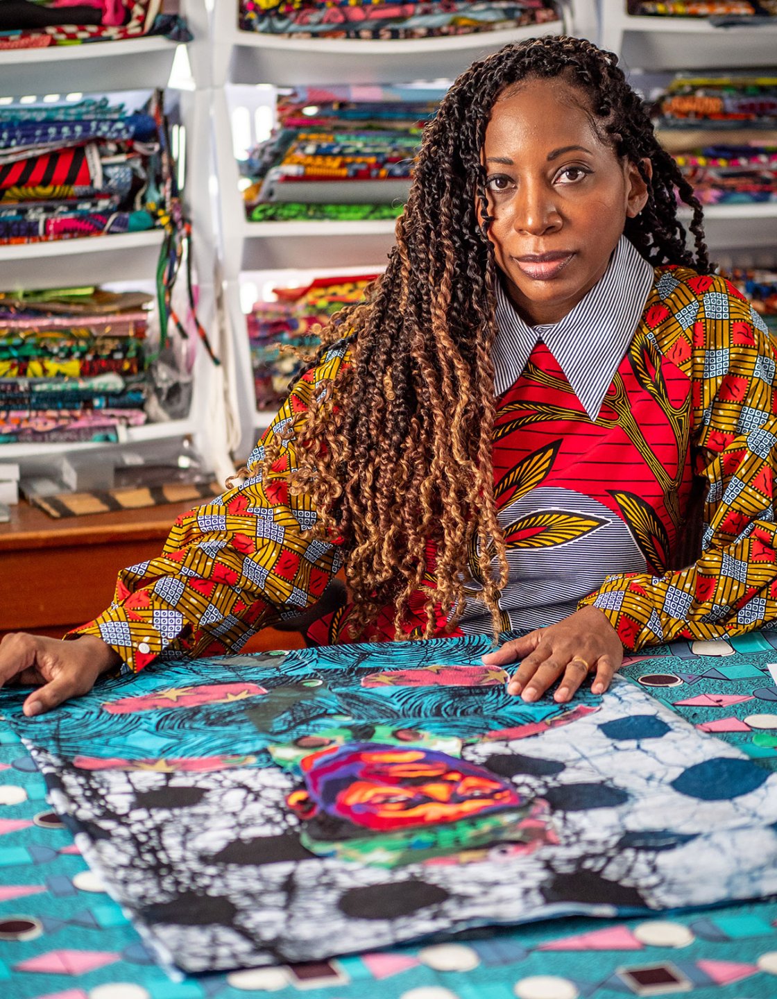 Bisa Butler seated at table with quilt in front of her and wall of fabrics behind