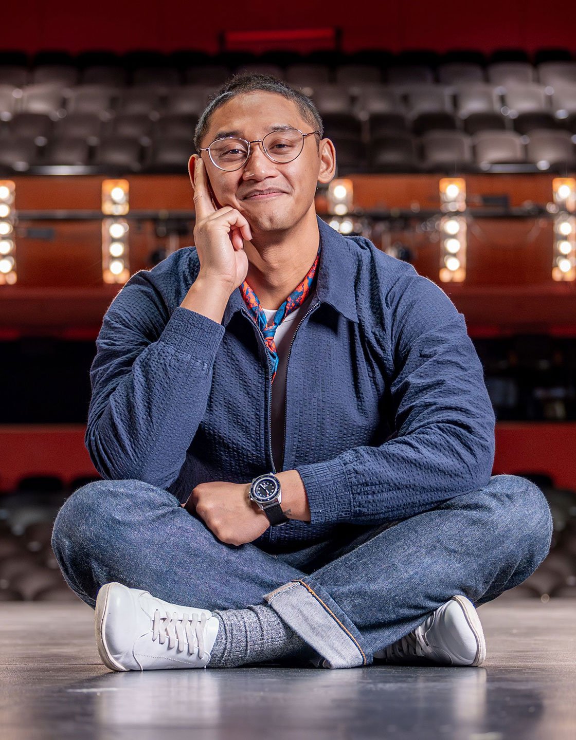 Josh De La Cruz sitting cross-legged on a stage in a theater