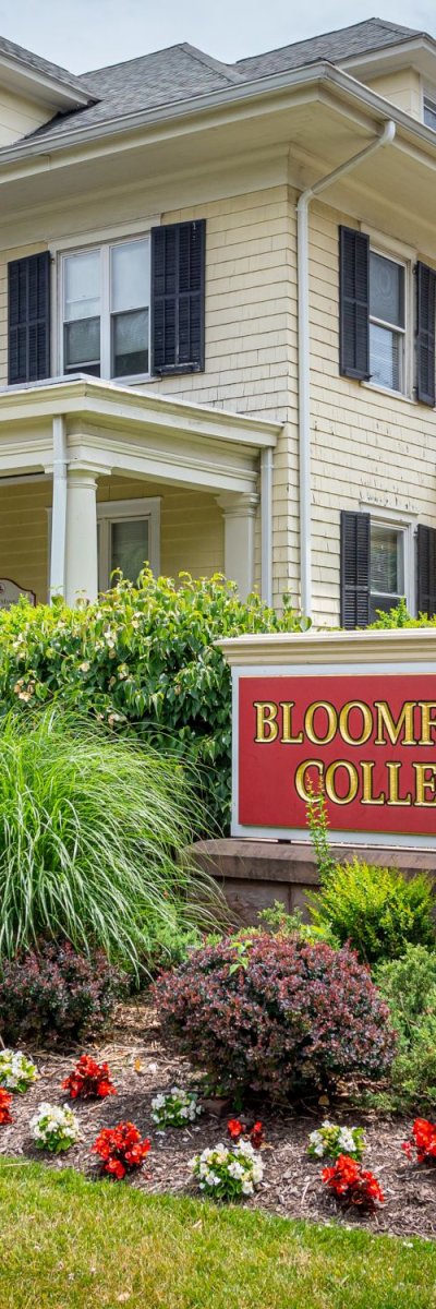 A yellow house with a Bloomfield College sign surrounded by green bushes.