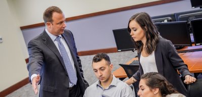Three students and a professor all in businesswear collaborate on a project unseen behind a multiple-monitor workstation
