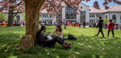 Two students relax under a flowering tree eating snacks from around the world while students from assorted campus culture clubs present for World's Fair Day in the background