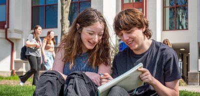 Two students sitting on the green grass of the Student Center Quad sharing a notebook