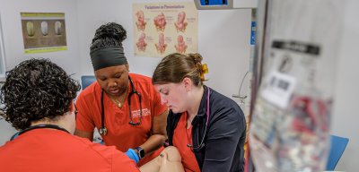 Three nursing students wearing red scrubs in a simulation lab tending to a manikin