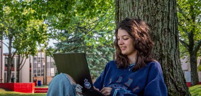 Student sitting under a tree, using their laptop.