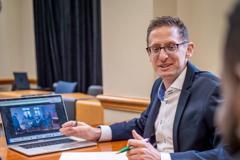Man is facing the camera explaining while his hand points to his computer.
