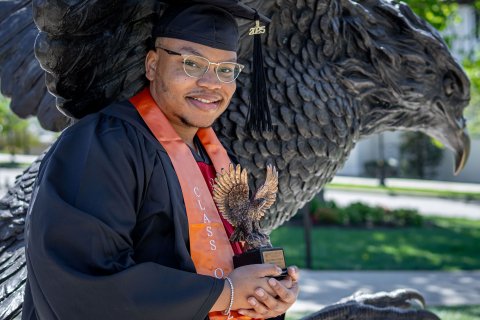 Emmanuel Ozias poses in cap and gown in front of the Red Hawk statute holding a trophy shaped like a red hawk.