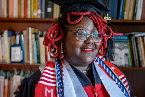 Khara Brown wears her cap and gown for a photograph taken next to a bookshelf inside Coder House, home to the Center for Heritage and Archaeological Studies.