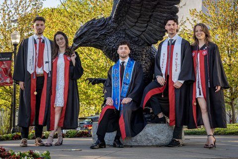 The Povolo quintuplets in graduation gowns stand in front of a large Red Hawk statue.